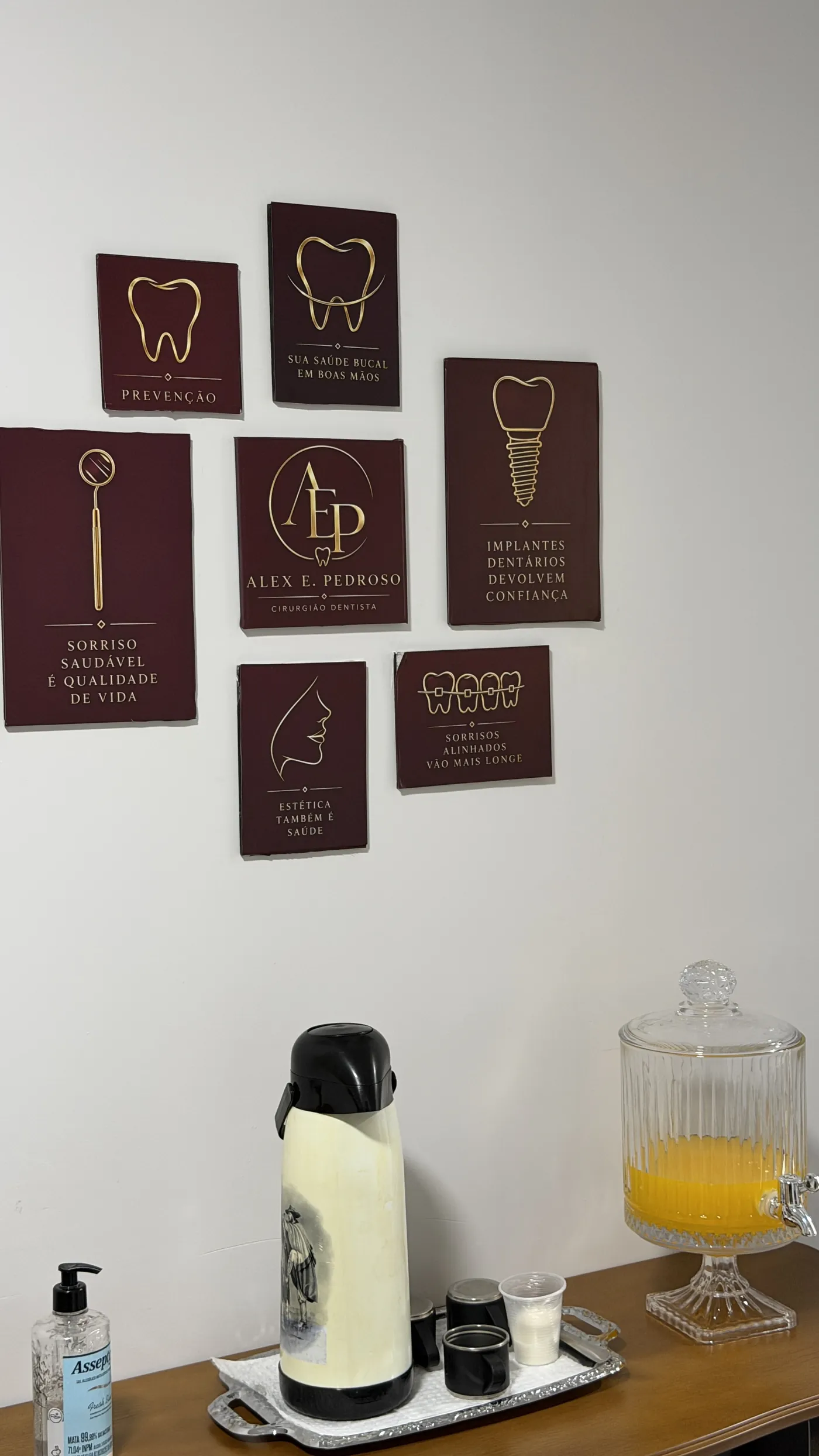Parede da recepção com quadros bordô e dourados da identidade do consultório e mesa de boas-vindas com café e suco